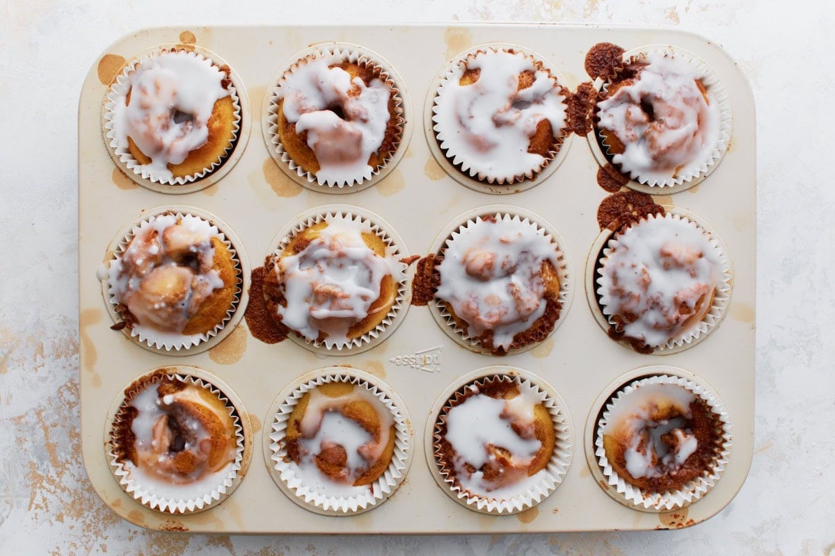 Overhead view of a muffin tin filled with freshly baked gluten-free cinnamon roll muffins, each topped with a generous drizzle of vanilla glaze.