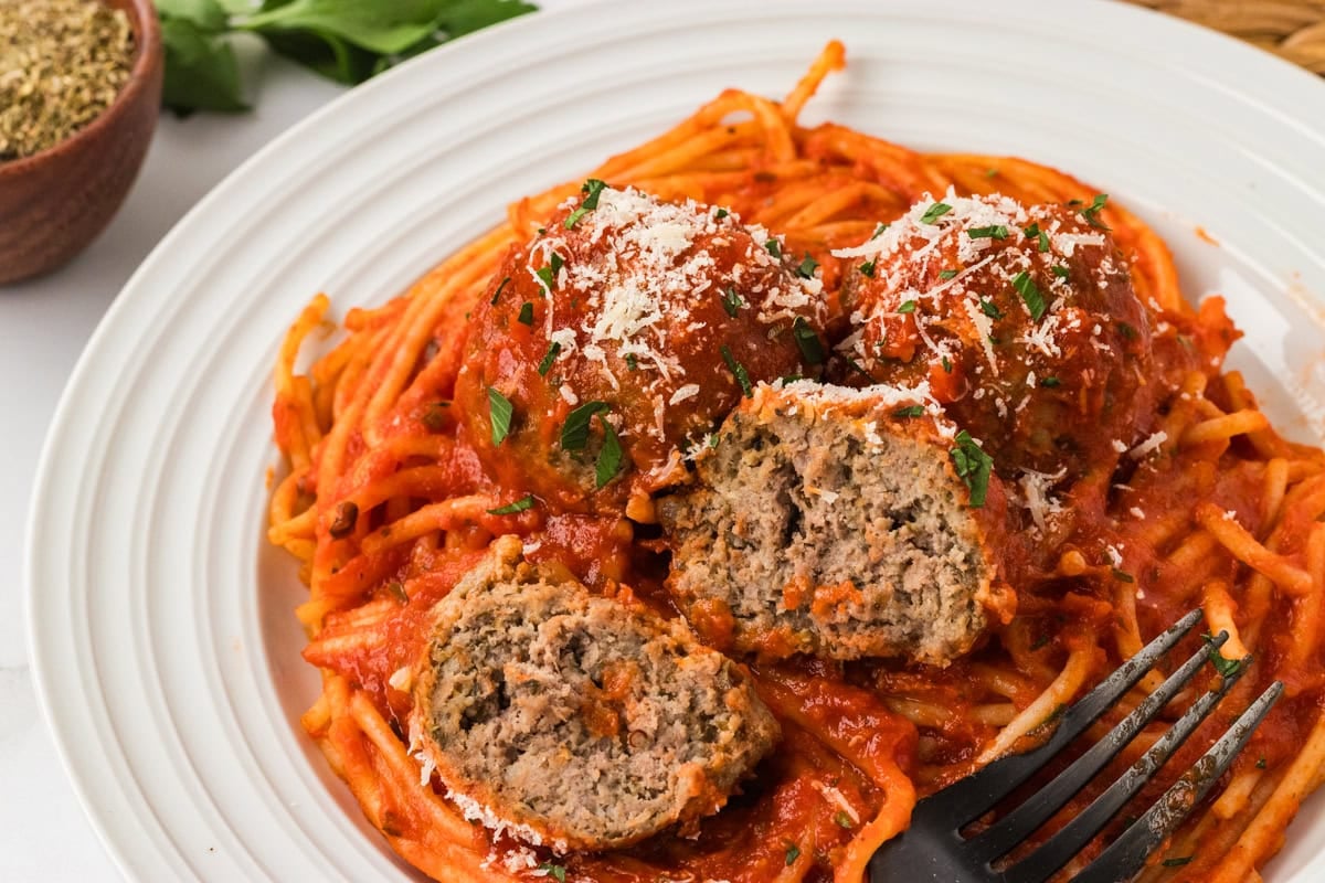 A plate of spaghetti topped with marinara sauce and three gluten-free meatballs, one cut open to show the juicy, tender center. Sprinkled with grated Parmesan and chopped parsley.