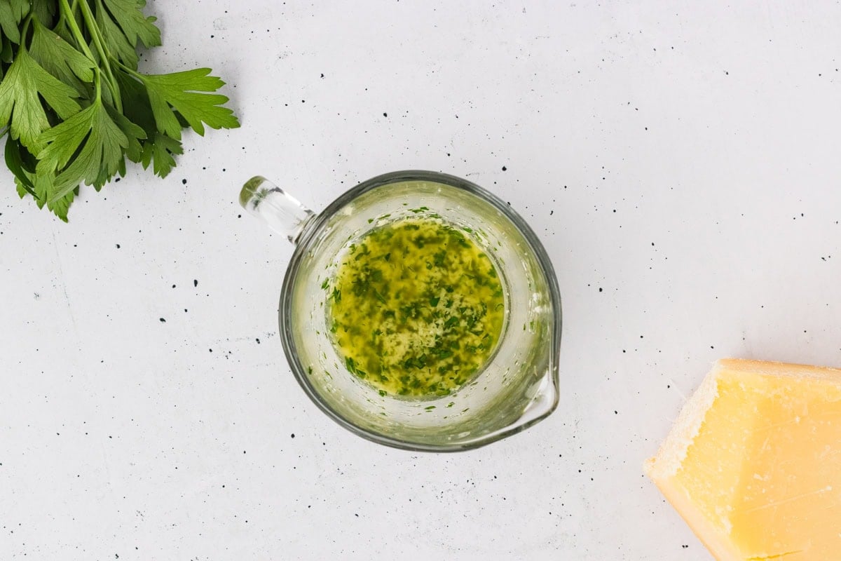 Small glass measuring cup filled with melted garlic herb butter, next to fresh parsley and a wedge of Parmesan.
