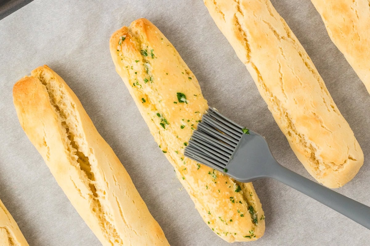 Silicone brush applying garlic herb butter onto warm gluten-free breadsticks.