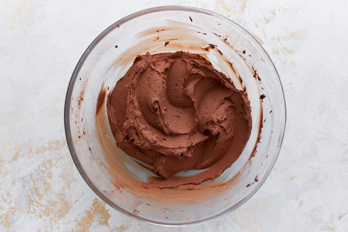 Thick, chocolate frosting forming as powdered sugar is added, shown in a mixing bowl with chocolate streaks.