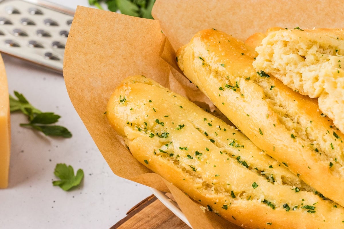 Golden, buttery gluten-free breadsticks in a parchment-lined bowl, one broken open to show the soft texture inside.