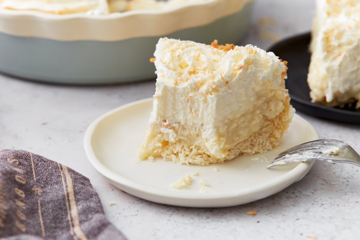 Close-up of a single slice of coconut cream pie on a white plate, with toasted coconut on top and a fork nearby.