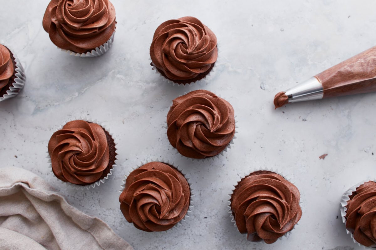 Overhead shot of frosted gluten-free chocolate cupcakes on a marble surface, with a piping bag to the side.