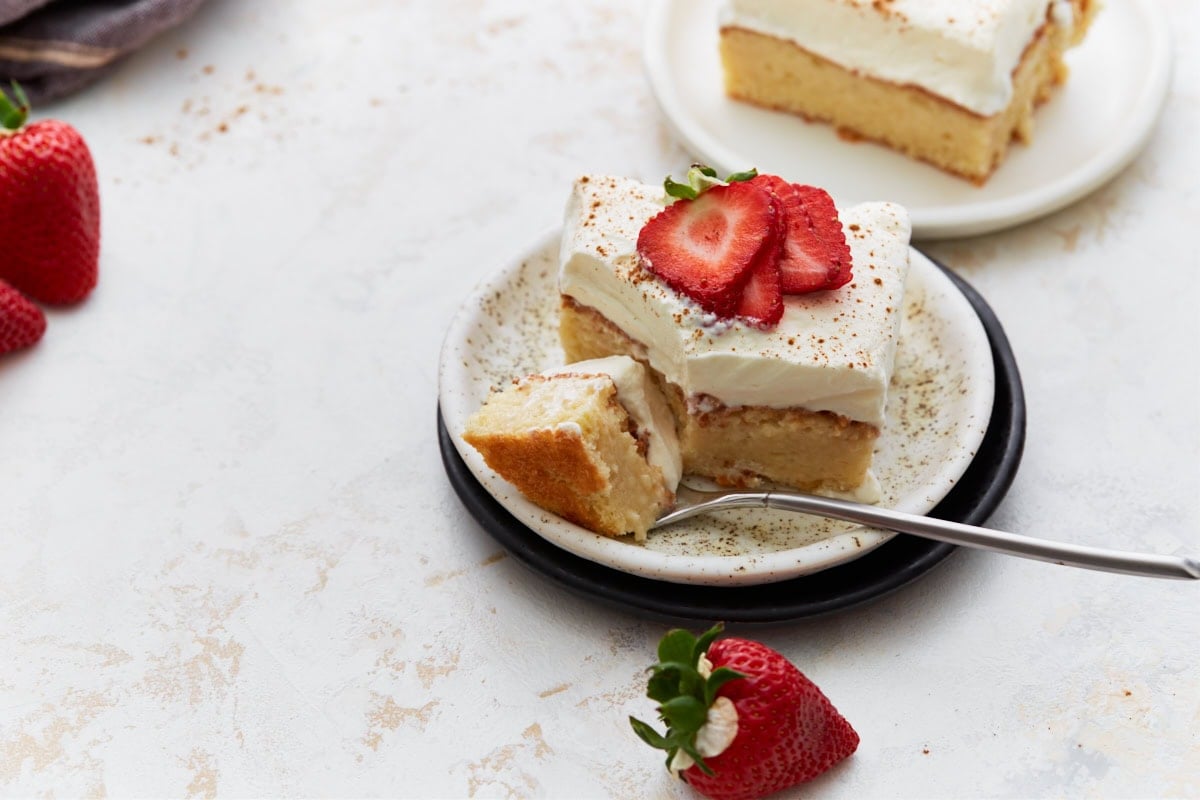 A slice of gluten-free tres leches cake topped with whipped cream and strawberries, served on a plate with a bite taken out on a fork, surrounded by fresh strawberries.