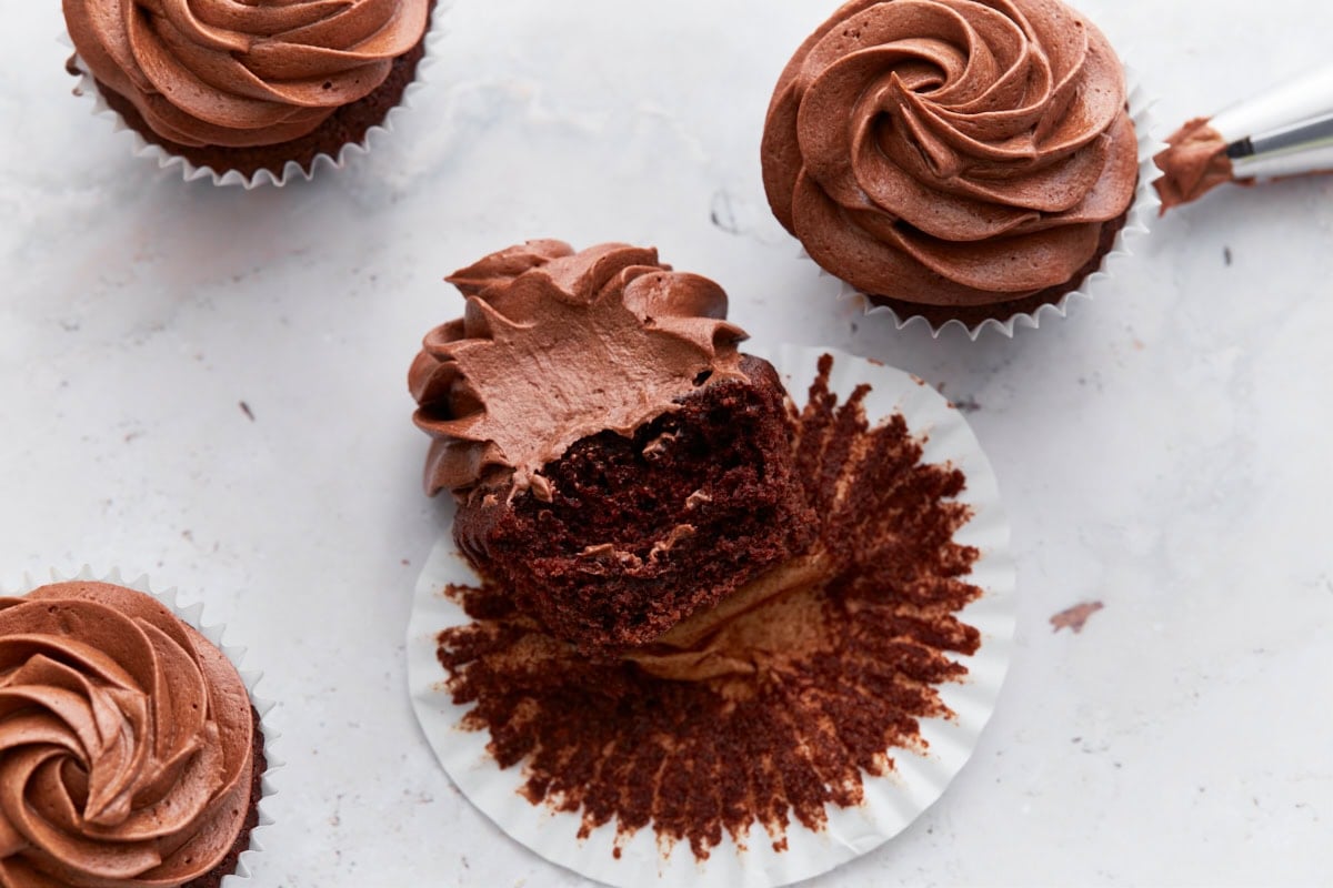 Close-up of a chocolate cupcake with a bite taken out, revealing a moist interior and fluffy chocolate frosting, surrounded by more frosted cupcakes.