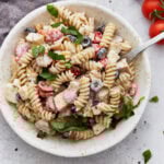 A bowl of creamy Italian pasta salad topped with fresh basil, filled with rotini pasta, cherry tomatoes, black olives, mozzarella cubes, and creamy dressing.