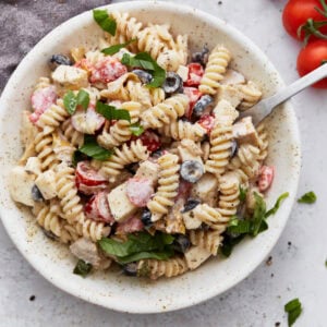 A bowl of creamy Italian pasta salad topped with fresh basil, filled with rotini pasta, cherry tomatoes, black olives, mozzarella cubes, and creamy dressing.