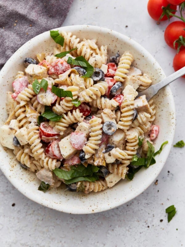 A bowl of creamy Italian pasta salad topped with fresh basil, filled with rotini pasta, cherry tomatoes, black olives, mozzarella cubes, and creamy dressing.