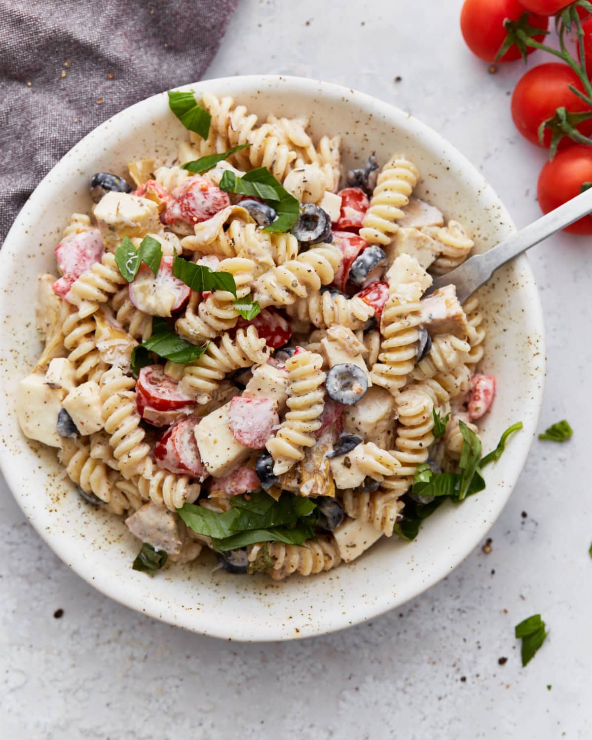 A bowl of creamy Italian pasta salad topped with fresh basil, filled with rotini pasta, cherry tomatoes, black olives, mozzarella cubes, and creamy dressing.