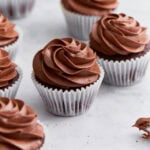 Close-up of several gluten-free chocolate cupcakes topped with swirled chocolate buttercream frosting in white cupcake liners on a white background.