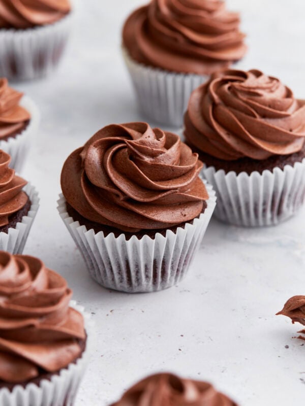Close-up of several gluten-free chocolate cupcakes topped with swirled chocolate buttercream frosting in white cupcake liners on a white background.