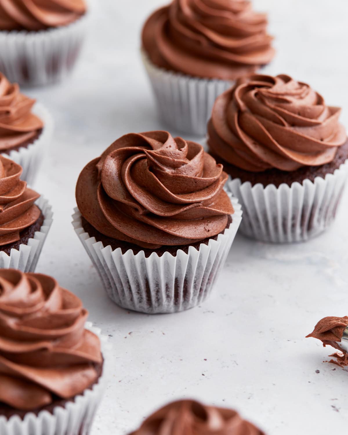Close-up of several gluten-free chocolate cupcakes topped with swirled chocolate buttercream frosting in white cupcake liners on a white background.