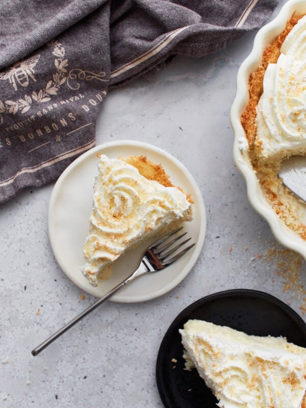 Slice of gluten-free coconut cream pie on a white plate with a fork, next to a full pie topped with whipped cream and toasted coconut.