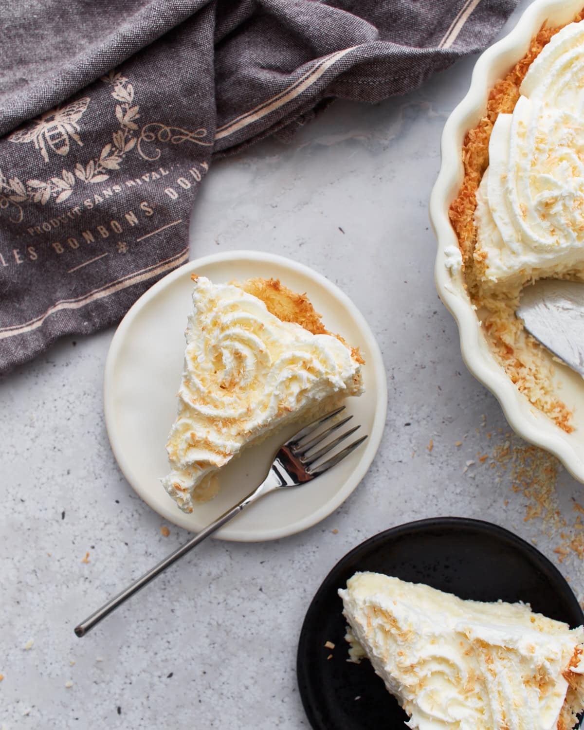 Slice of gluten-free coconut cream pie on a white plate with a fork, next to a full pie topped with whipped cream and toasted coconut.