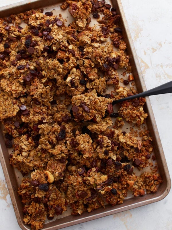 A close-up of a baking sheet filled with chunky, golden-brown gluten-free granola featuring visible chocolate chips, raisins, and mixed nuts, with a black spoon scooping some up.