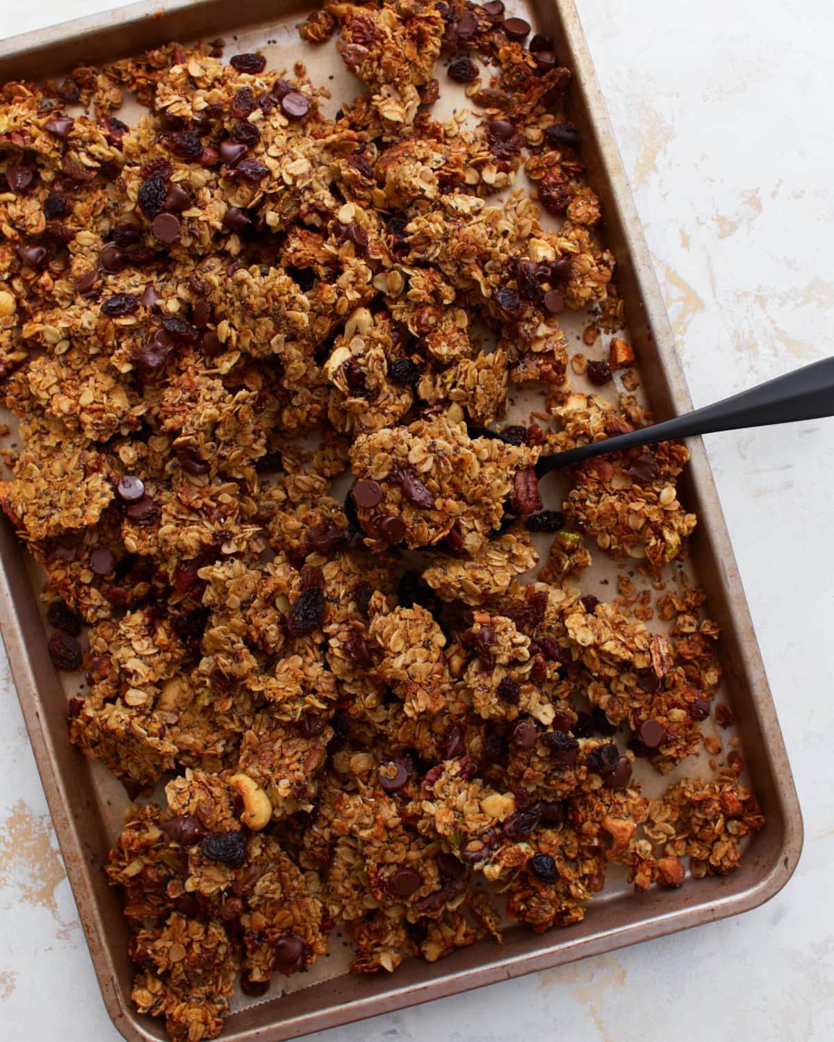 A close-up of a baking sheet filled with chunky, golden-brown gluten-free granola featuring visible chocolate chips, raisins, and mixed nuts, with a black spoon scooping some up.