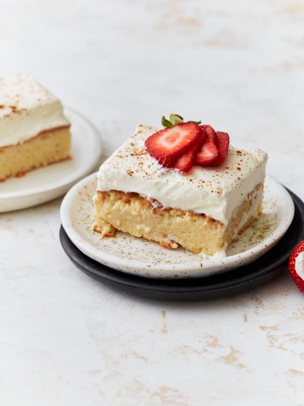 A slice of gluten-free tres leches cake topped with whipped cream and fresh strawberry slices, served on a speckled plate.