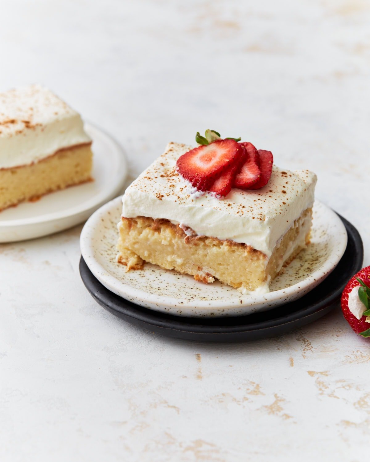 A slice of gluten-free tres leches cake topped with whipped cream and fresh strawberry slices, served on a speckled plate.