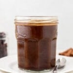 A glass jar filled with dark brown homemade Worcestershire sauce, placed on a stack of white plates with a spoon beside it.