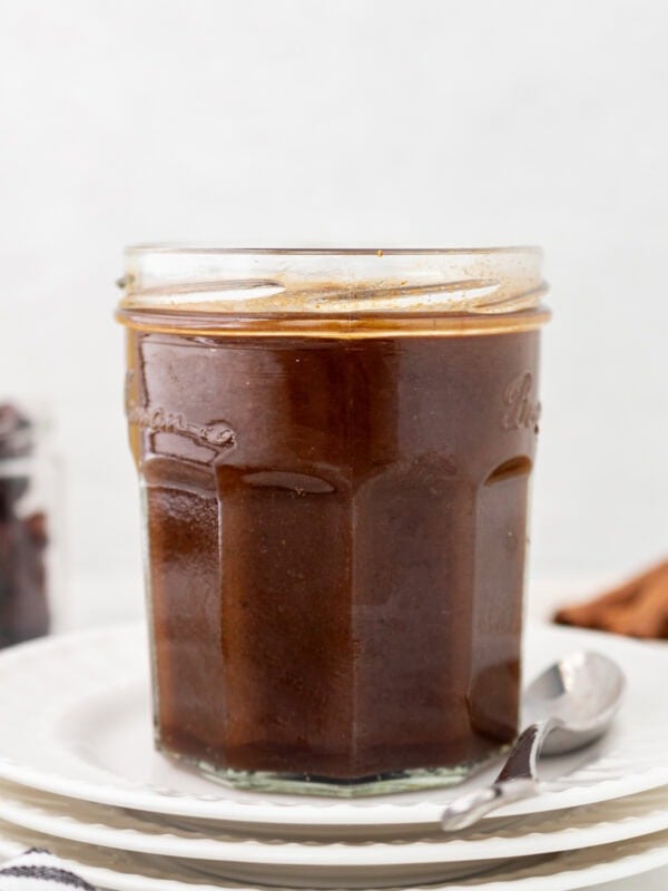 A glass jar filled with dark brown homemade Worcestershire sauce, placed on a stack of white plates with a spoon beside it.