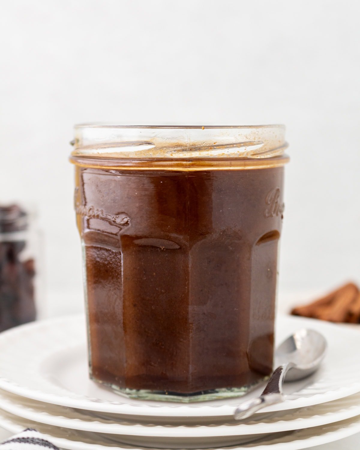 A glass jar filled with dark brown homemade Worcestershire sauce, placed on a stack of white plates with a spoon beside it.