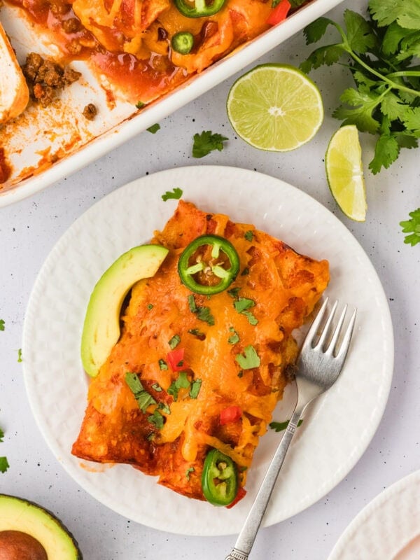 A serving of cheesy gluten-free beef and cheese enchiladas on a white plate, topped with jalapeño slices, avocado, and chopped cilantro, with a baking dish and lime wedges in the background.