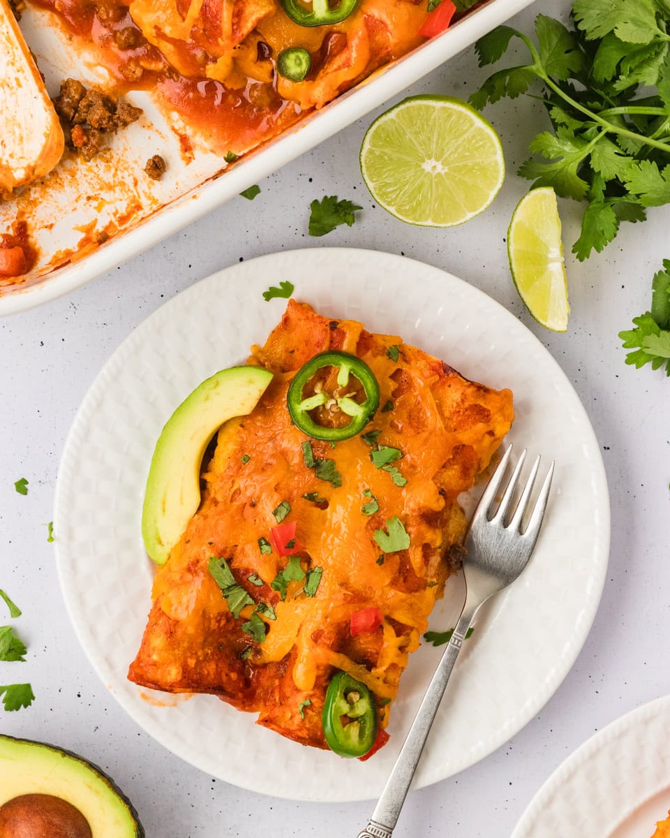 A serving of cheesy gluten-free beef and cheese enchiladas on a white plate, topped with jalapeño slices, avocado, and chopped cilantro, with a baking dish and lime wedges in the background.
