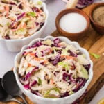A close-up of two white scalloped bowls filled with colorful dairy-free coleslaw, placed on a wooden board with small bowls of salt and pepper and a halved red cabbage in the background.