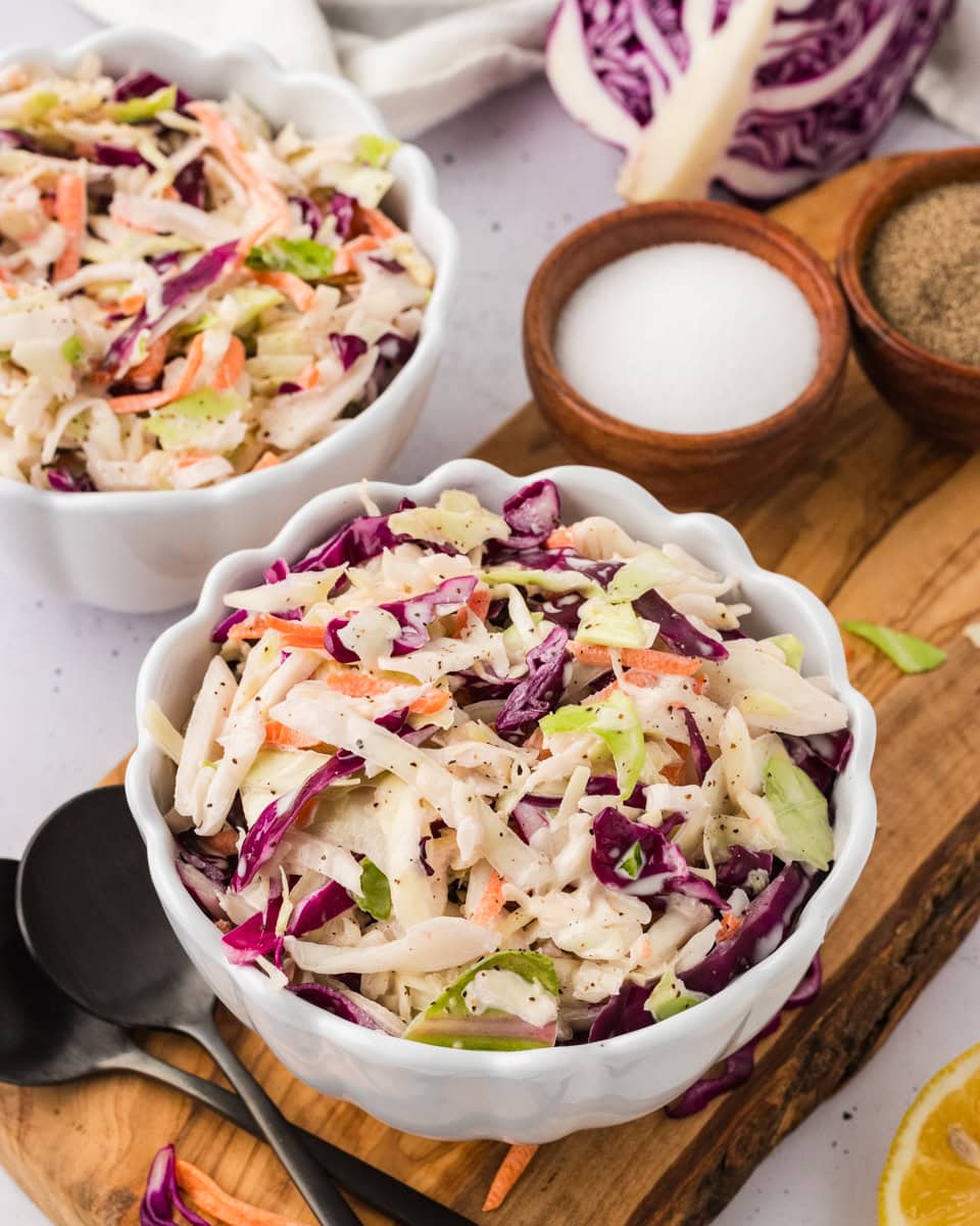 A close-up of two white scalloped bowls filled with colorful dairy-free coleslaw, placed on a wooden board with small bowls of salt and pepper and a halved red cabbage in the background.
