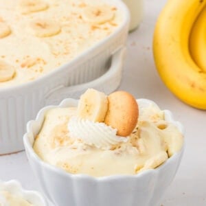 Close-up of a serving of gluten-free banana pudding in a white scalloped bowl, topped with whipped cream, a banana slice, and a gluten-free vanilla wafer cookie. A large dish of banana pudding and whole bananas are in the background.