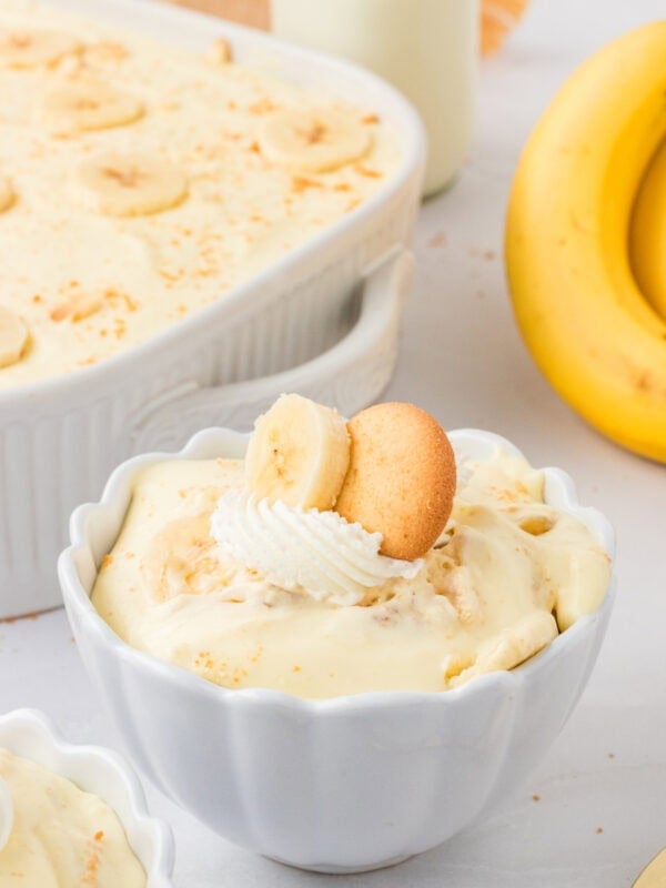 Close-up of a serving of gluten-free banana pudding in a white scalloped bowl, topped with whipped cream, a banana slice, and a gluten-free vanilla wafer cookie. A large dish of banana pudding and whole bananas are in the background.