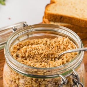 Close-up of a glass jar filled with golden gluten-free breadcrumbs, with a metal spoon inside and slices of gluten-free bread in the background.