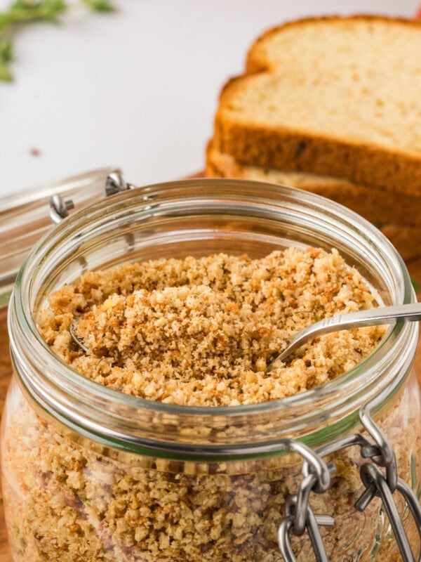 Close-up of a glass jar filled with golden gluten-free breadcrumbs, with a metal spoon inside and slices of gluten-free bread in the background.
