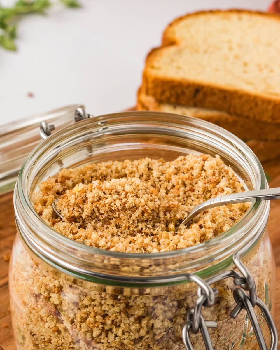 Close-up of a glass jar filled with golden gluten-free breadcrumbs, with a metal spoon inside and slices of gluten-free bread in the background.