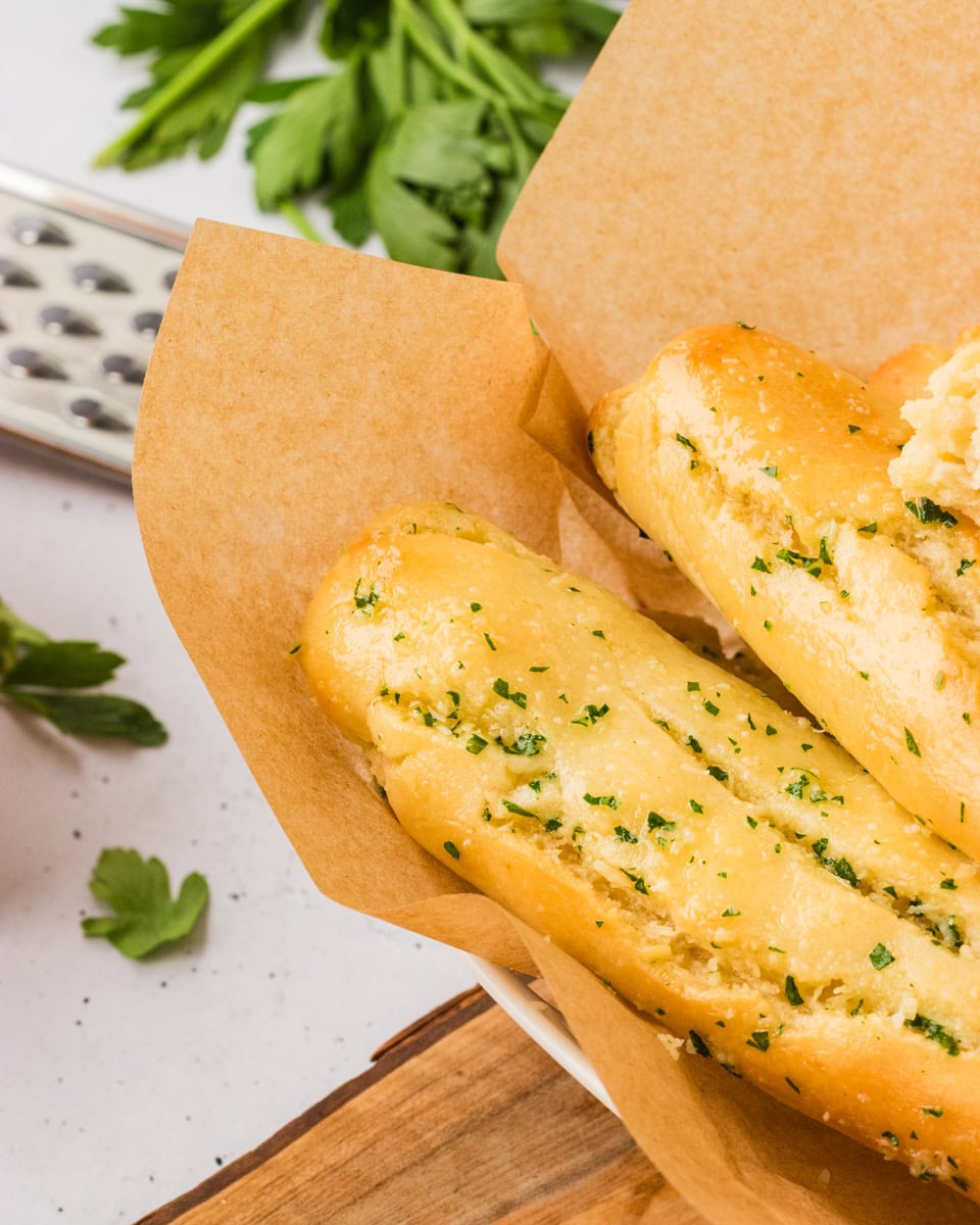 Close-up of golden gluten-free breadsticks topped with garlic and parsley, served in a parchment-lined bowl.