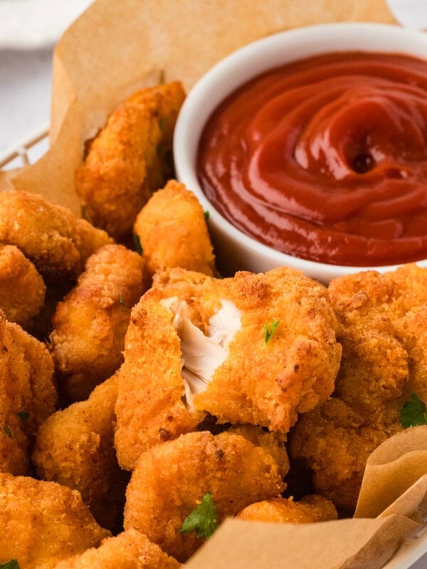Close-up of crispy gluten-free chicken nuggets in a basket with parchment paper, served with a bowl of ketchup for dipping. One nugget is broken open to show the juicy inside.