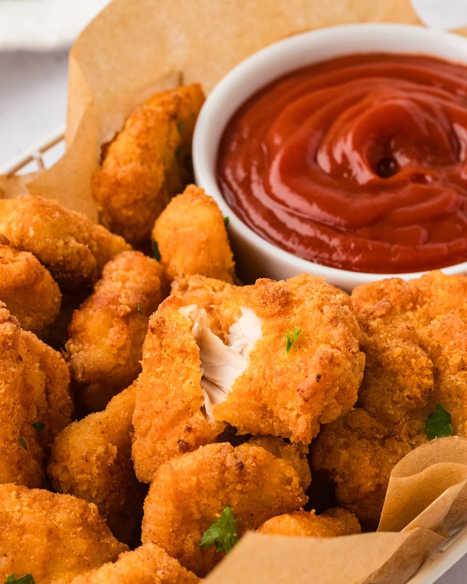 Close-up of crispy gluten-free chicken nuggets in a basket with parchment paper, served with a bowl of ketchup for dipping. One nugget is broken open to show the juicy inside.