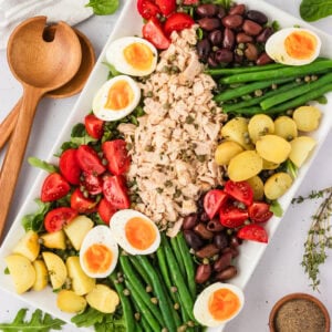 Overhead view of a beautifully arranged Tuna Nicoise Salad on a white rectangular platter with eggs, tomatoes, olives, green beans, potatoes, and tuna.