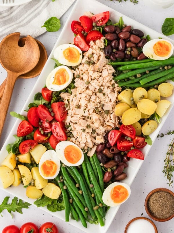 Overhead view of a beautifully arranged Tuna Nicoise Salad on a white rectangular platter with eggs, tomatoes, olives, green beans, potatoes, and tuna.