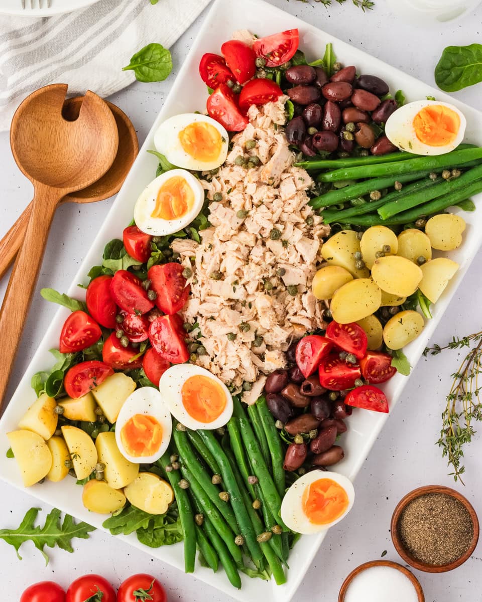 Overhead view of a beautifully arranged Tuna Nicoise Salad on a white rectangular platter with eggs, tomatoes, olives, green beans, potatoes, and tuna.