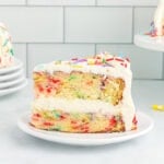 A close-up of a slice of gluten-free funfetti cake with white buttercream frosting and rainbow sprinkles, showing the colorful sprinkle-filled layers inside.