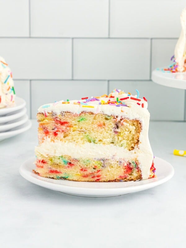 A close-up of a slice of gluten-free funfetti cake with white buttercream frosting and rainbow sprinkles, showing the colorful sprinkle-filled layers inside.
