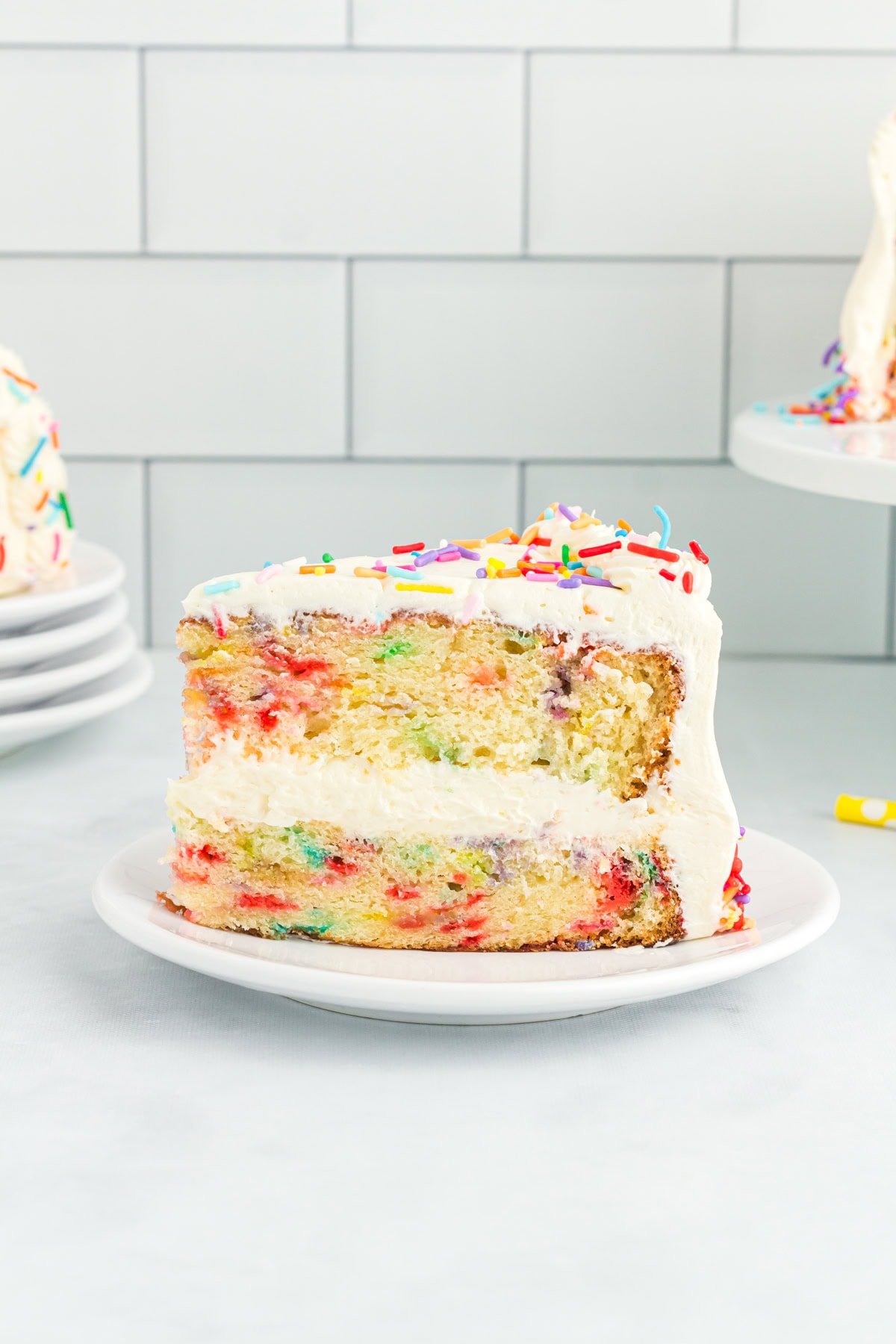 A close-up of a slice of gluten-free funfetti cake with white buttercream frosting and rainbow sprinkles, showing the colorful sprinkle-filled layers inside.