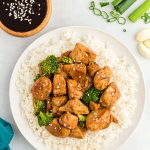 Plate of chicken and broccoli stir-fry served over white rice, garnished with sesame seeds, with a bowl of hoisin sauce, green onions, and garlic cloves on the side.