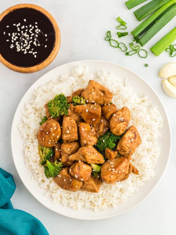 Plate of chicken and broccoli stir-fry served over white rice, garnished with sesame seeds, with a bowl of hoisin sauce, green onions, and garlic cloves on the side.