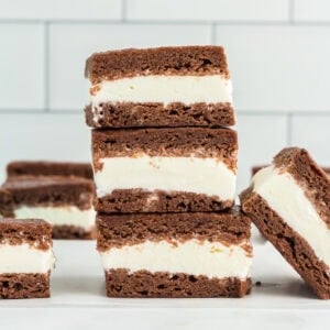 Stack of gluten-free ice cream sandwiches with creamy vanilla filling between two layers of chocolate cookie, set against a white tiled background.
