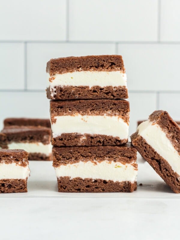 Stack of gluten-free ice cream sandwiches with creamy vanilla filling between two layers of chocolate cookie, set against a white tiled background.