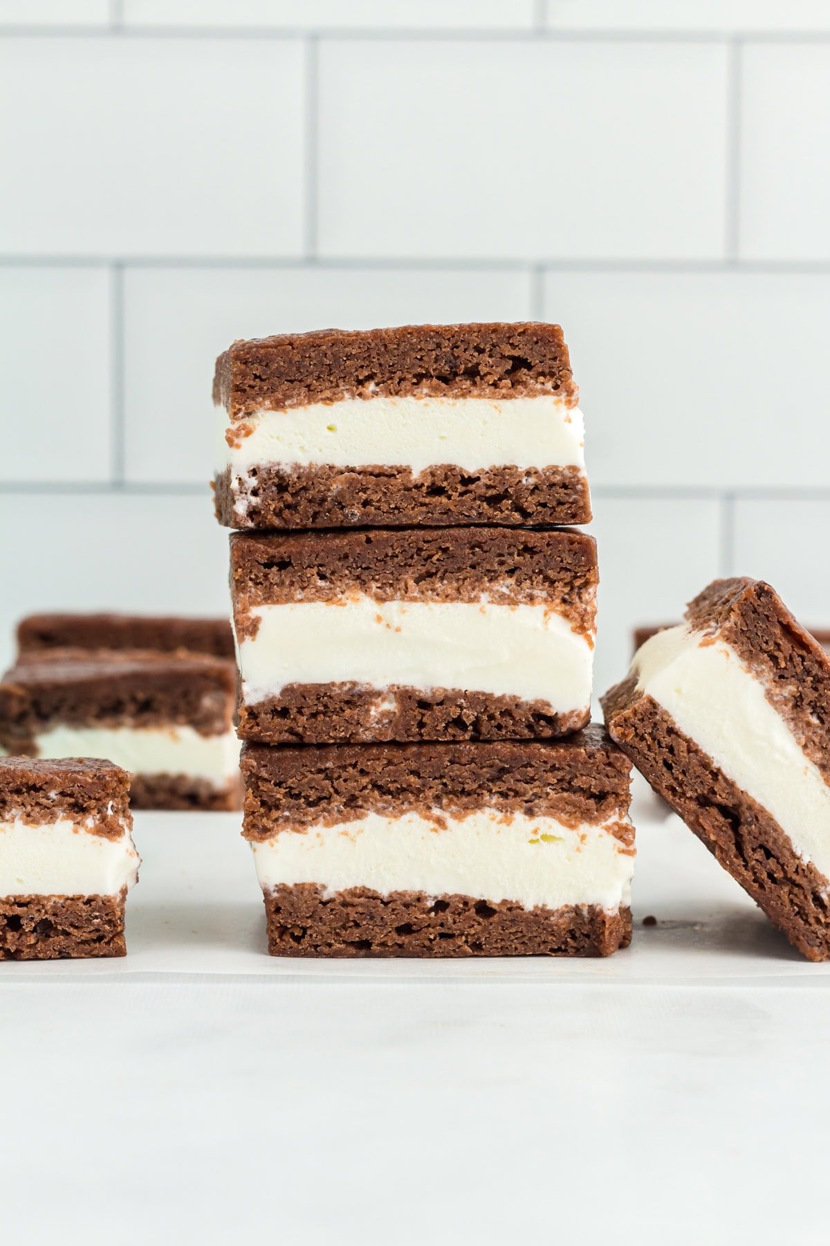 Stack of gluten-free ice cream sandwiches with creamy vanilla filling between two layers of chocolate cookie, set against a white tiled background.