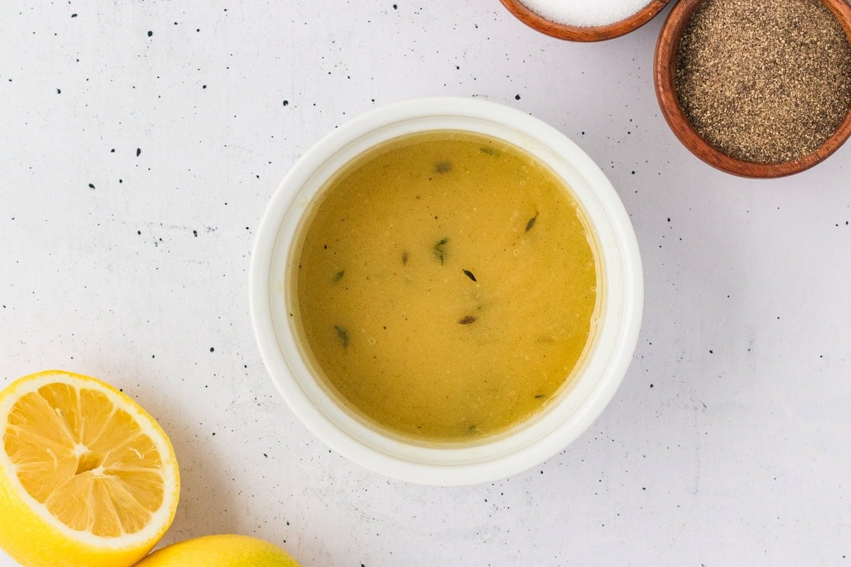 mall white bowl of lemon vinaigrette surrounded by bowls of salt, pepper, and fresh lemons on a light background.